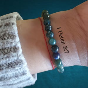 May include: A beaded bracelet featuring smooth, round, multi-colored stones in shades of deep green, blue, and dark teal. A thin red string bracelet is worn alongside it. The text 1 Peter 5:7 is visible on the skin next to the beaded bracelet.