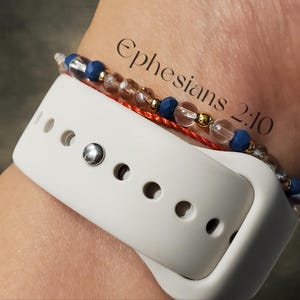 May include: A white Apple Watch band with a red string and a beaded bracelet with blue and clear beads. The bracelet has gold accents and the text "Ephesians 2:10" written on the wrist.