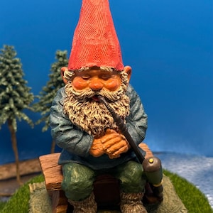 Könnte beinhalten: Detaillierte Figur eines Gartenzwergs, der einen roten Spitzhut, eine blaue Jacke und eine grüne Hose trägt. Der Zwerg hat einen langen, weißen Bart und raucht eine Pfeife, während er auf einer Holzbank sitzt. Die Figur steht auf einem grünen Sockel.