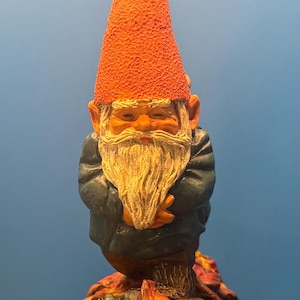 Könnte beinhalten: Eine Gartenzwerg-Statue mit einem hohen, strukturierten orangefarbenen Hut und einem langen, weißen Bart. Der Zwerg trägt einen dunkelblauen Mantel und hat die Arme verschränkt. Der Sockel zeigt bunte Herbstblätter und steht auf grünem Gras.