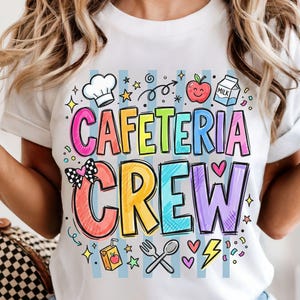 May include: White t-shirt with the words "CAFETERIA CREW" in colorful, bubble letters. The design includes a chef's hat, apple, milk carton, and other food-related graphics. The shirt has short sleeves.