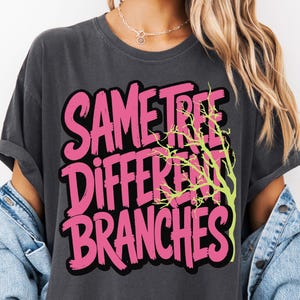 May include: Dark grey t-shirt featuring the text "SAME TREE DIFFERENT BRANCHES" in pink with a black outline. A lime green tree graphic is on the right. The shirt has rolled-up sleeves.