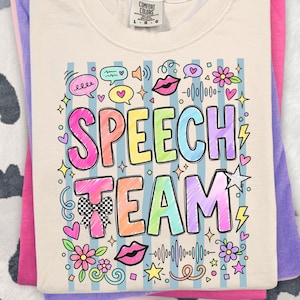 May include: Cream-coloured t-shirt with the words "SPEECH TEAM" in colourful, bubble letters. The design includes speech bubbles, hearts, flowers, and sound wave graphics. The shirt is folded with other shirts in shades of pink and purple.