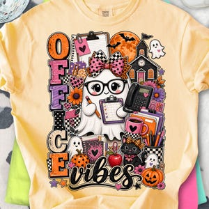 Peut inclure: Un t-shirt jaune pâle avec une impression graphique représentant un fantôme portant des lunettes et un nœud à motif léopard, tenant un presse-papier et un crayon. Le motif inclut le texte "OFFICE vibes", un téléphone noir, un chat et des citrouilles.