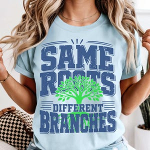 May include: Pale blue t-shirt with the slogan "SAME ROOTS DIFFERENT BRANCHES" in blue and green. The design includes a tree graphic. The shirt has short sleeves and a round neck.