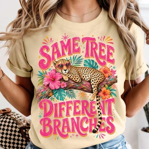 May include: A light yellow t-shirt with the text "SAME TREE DIFFERENT BRANCHES" in pink. The design includes a cheetah in sunglasses on a branch with flowers. The shirt has rolled-up sleeves.