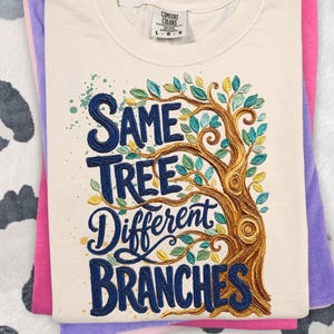 May include: Cream-coloured t-shirt with the words "Same Tree Different Branches" in navy blue script, featuring a detailed tree illustration with green and yellow leaves. The shirt is folded on top of pink and purple shirts.