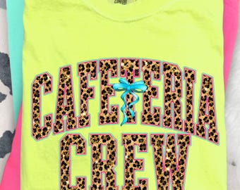 May include: Neon yellow t-shirt with the words "CAFETERIA CREW" in a leopard print design, accented with a light blue bow. The shirt is stacked with other brightly colored shirts.