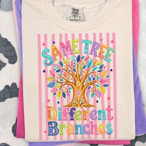May include: Cream-coloured t-shirt featuring a colourful tree graphic and the text "Same Tree Different Branches." The tree has a brown trunk and branches with leaves in various colours. The background has pink stripes and stars.