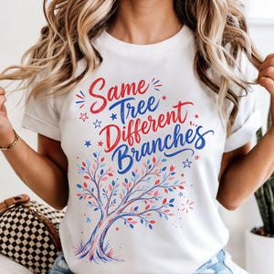 May include: White t-shirt featuring the text "Same Tree Different Branches" in red and blue. The design includes a tree with red, blue, and pink leaves. The shirt is short-sleeved.