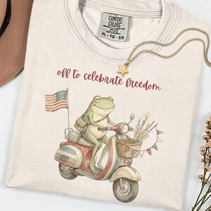 Könnte beinhalten: Cremefarbenes T-Shirt mit einer Frosch-Illustration auf einem rot-weiß-blauen Roller, der eine amerikanische Flagge und Feuerwerk trägt. Der Text "off to celebrate freedom" ist rot gedruckt. Eine goldene Sternkette liegt auf dem Shirt.