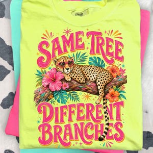 May include: Neon yellow t-shirt featuring a cheetah wearing sunglasses, lounging on a branch with flowers. The text "SAME TREE DIFFERENT BRANCHES" is in pink and orange. The shirt is stacked with pink and teal shirts.