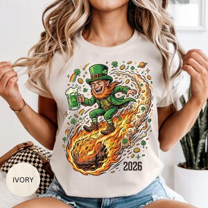May include: Ivory-colored t-shirt featuring a cartoon leprechaun riding a fiery comet, holding a beer mug with a shamrock. The design includes rainbows, gold coins, and the year "2026". The shirt is a festive design for St. Patrick's Day.