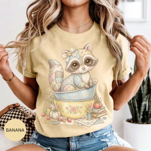 Könnte beinhalten: Hellgelbes T-Shirt mit einem Cartoon-Waschbären in einer Schüssel. Der Waschbär hat einen Flickenpelz und eine Schleife. Die Schüssel hat florale Muster und steht auf einer anderen Schüssel. Nähutensilien und Bücher sind ebenfalls zu sehen.