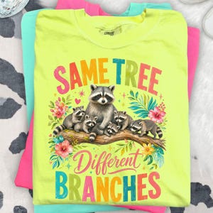 May include: A neon yellow t-shirt with a raccoon family illustration, floral accents, and the text "SAME TREE Different BRANCHES". The shirt is layered with pink and turquoise garments.