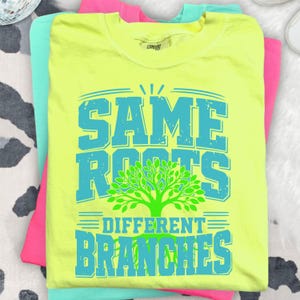 May include: A neon yellow t-shirt with the text "SAME ROOTS DIFFERENT BRANCHES" in turquoise. A green tree graphic is centered below the words "ROOTS". The shirt is stacked with other brightly coloured shirts.