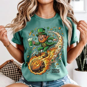 May include: Teal green t-shirt featuring a cartoon leprechaun riding a fiery comet, holding a beer mug. The design includes shamrocks, rainbows, and the year "2026". A festive St. Patrick's Day themed graphic.