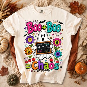 May include: A cream-coloured t-shirt featuring a colourful graphic of a ghost holding a chalkboard with math equations. The design includes the text Boo + Boo = Chaos, surrounded by flowers, small red apples, stars, and bats in vibrant colours.