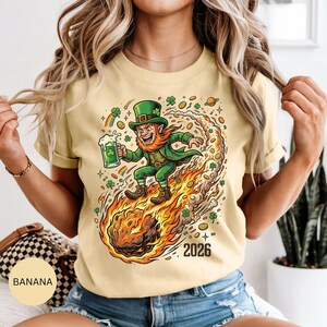 May include: A light yellow t-shirt featuring a cartoon leprechaun riding a fiery comet, holding a green beer. The leprechaun wears a green hat and coat. The shirt has the text "2026" and is decorated with shamrocks and rainbows.