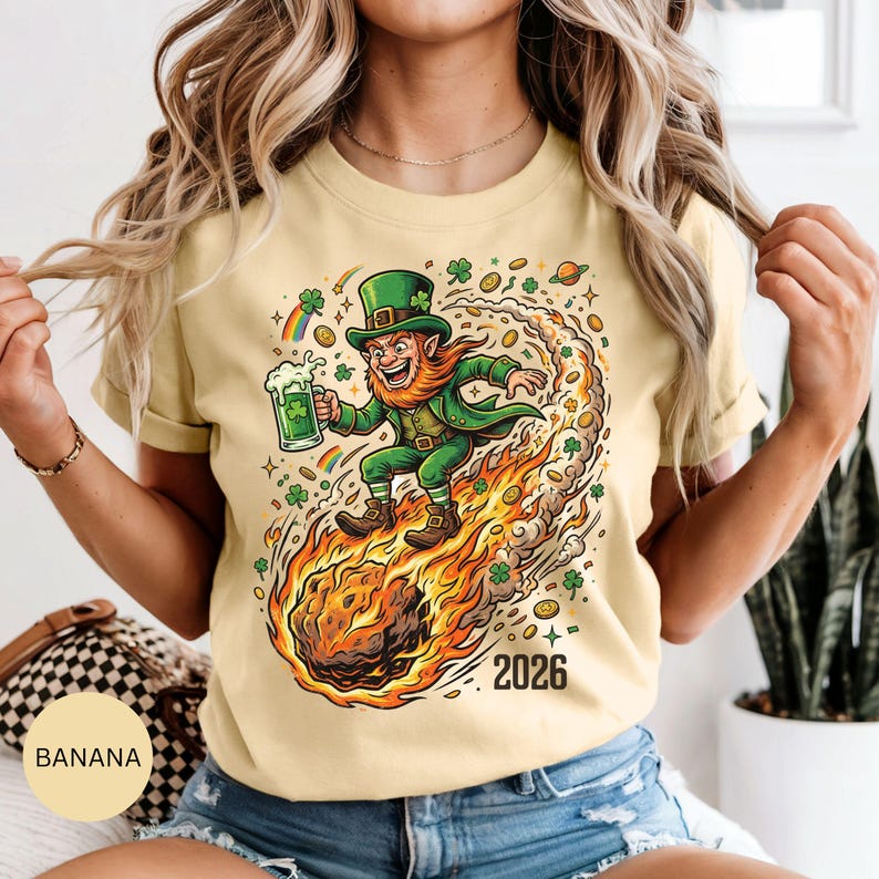 May include: A light yellow t-shirt featuring a cartoon leprechaun riding a fiery comet, holding a green beer. The leprechaun wears a green hat and coat. The shirt has the text "2026" and is decorated with shamrocks and rainbows.
