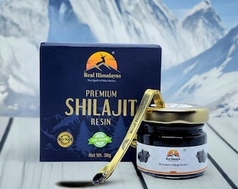 Puede incluir: Una caja azul marino y un pequeño tarro de cristal con tapa dorada, ambos etiquetados como "Premium Shilajit Resin". Una cuchara dorada descansa junto a ellos. La caja tiene el texto "Real Himalayas" y "Net Wt. 30g."