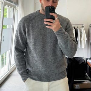 Peut inclure: Un pull à manches longues gris chiné doté d'une encolure ronde ainsi que de poignets et d'un ourlet en tricot côtelé. Le vêtement est conçu avec des manches raglan et un motif en tricot texturé, offrant une teinte grise uniforme sur l'ensemble.