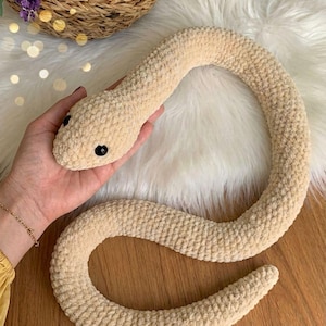 Puede incluir: Un juguete de serpiente de ganchillo beige con ojos negros. La serpiente de peluche está hecha de un hilo suave y texturizado y está enrollada en forma circular. La serpiente se muestra sobre una superficie blanca y esponjosa.