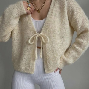 May include: A cream-coloured, long-sleeved, fuzzy knit cardigan featuring a V-neckline and a single tie-front closure. The cardigan is worn over a white ribbed tank top. The garment has a soft, textured appearance with ribbed cuffs and hem.