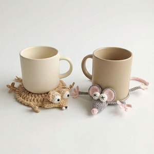 Könnte beinhalten: Zwei beige Keramikbecher stehen auf handgefertigten Häkeluntersetzern. Der eine Untersetzer ist ein hellbraunes Wesen mit großen Augen, der andere eine graue Maus mit rosa Ohren und Pfoten. Auf einer weißen Oberfläche.