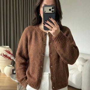 Pode incluir: Um cardigã de malha castanho com decote redondo, mangas compridas com punhos canelados e fecho frontal de botões. A peça é usada sobre uma blusa branca lisa e calças brancas, exibindo um padrão de malha texturizada e um corte padrão.