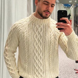 May include: A cream-colored cable knit sweater featuring a ribbed mock neck collar and long sleeves. The garment displays a vertical pattern of intertwined rope-like cables and textured stitches, providing a thick, warm appearance.