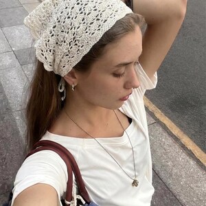 May include: A cream-colored crochet headscarf featuring an intricate openwork pattern, worn tied at the back. The accessory is paired with a plain white short-sleeved t-shirt, a delicate gold-tone chain necklace with a pendant, and small gold hoop earrings.