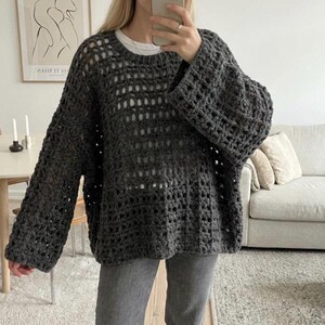 May include: A dark charcoal gray long-sleeve sweater featuring an open-knit crochet pattern with a wide, loose fit, a round neckline, and dropped shoulders, worn over a plain white t-shirt and paired with gray denim jeans.