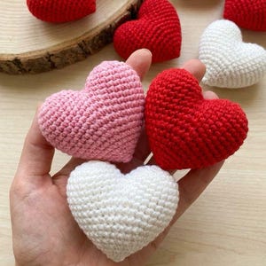 May include: A collection of handmade crochet hearts in various colors, including red, pink, and white. The hearts are displayed on a wooden surface, showcasing their textured, knitted appearance. These decorative hearts are perfect for Valentine's Day or other special occasions.