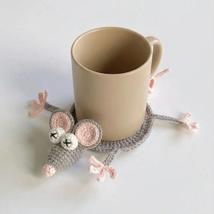 May include: A beige ceramic mug sits on a grey crocheted mouse-shaped coaster. The coaster has pink accents on the ears, nose, and paws, with white eyes and black cross details. The mug has a simple, cylindrical design.