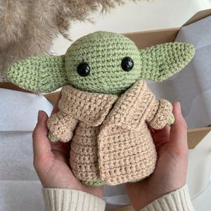 Op de afbeelding: Gehaakte pop van een personage met grote oren, groene huid en een beige gewaad. De pop heeft grote zwarte ogen. De pop wordt in iemands handen gehouden. De pop zit in een kartonnen doos.