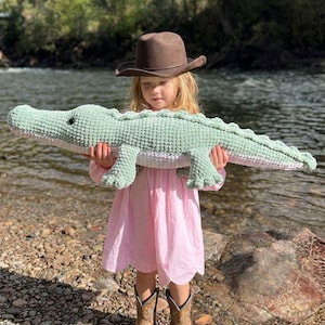 Puede incluir: Un juguete de peluche de cocodrilo tejido en color verde claro, sostenido por un niño con un vestido rosa y botas de vaquero. El niño también lleva un sombrero de vaquero marrón. El fondo muestra un río y árboles.
