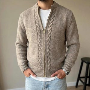 Peut inclure: Un gilet zippé en tricot beige avec un motif de torsades vertical le long de la fermeture éclair, des manches longues à poignets côtelés et un ourlet côtelé, porté sur un t-shirt blanc uni avec un jean bleu clair.