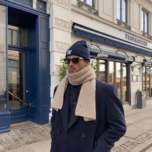 Può includere: Un berretto blu navy con una lettera W bianca sul davanti, occhiali da sole neri, una sciarpa in maglia a coste beige e un cappotto in lana doppiopetto blu navy. L'outfit è indossato sopra un gilet trapuntato scuro per un look a strati.