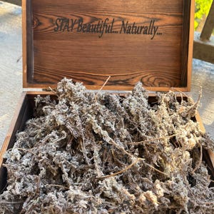 May include: A wooden box filled with dried herbs. The box is open, revealing a collection of dried, gray-green herbs. The box's lid has the text "STAY Beautiful... Naturally..." etched into the wood.