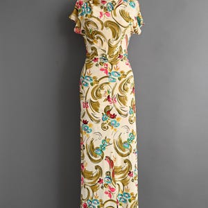 May include: A vintage cream-colored dress with a floral pattern. The dress features short sleeves and a fitted bodice that flows into a long skirt. The floral design includes olive green, pink, and blue accents. The dress is displayed on a mannequin.