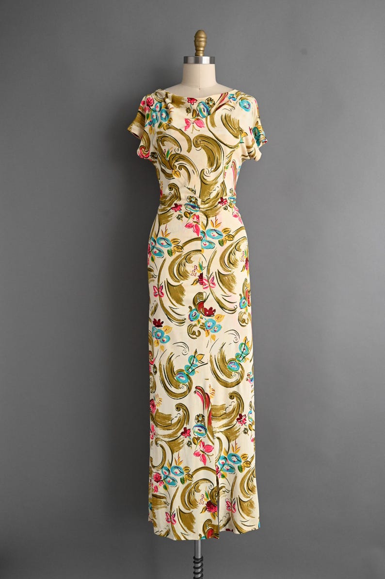 May include: A vintage cream-colored dress with a floral pattern. The dress features short sleeves and a fitted bodice that flows into a long skirt. The floral design includes olive green, pink, and blue accents. The dress is displayed on a mannequin.