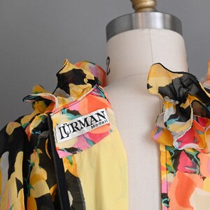 May include: A black and yellow floral print blouse with a ruffled collar and a zipper closure. The blouse has a yellow label with the text "LURMAN Modell" on the front.