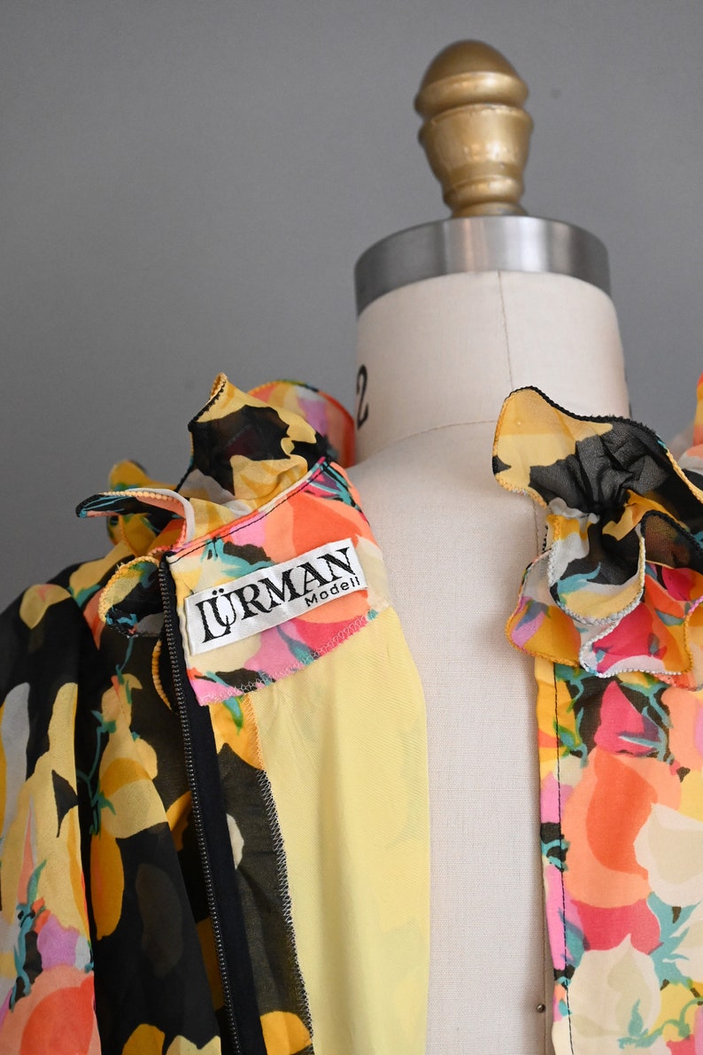 May include: A black and yellow floral print blouse with a ruffled collar and a zipper closure. The blouse has a yellow label with the text "LURMAN Modell" on the front.