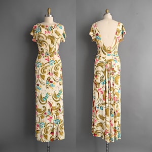 May include: A vintage cream-colored dress with a colorful floral pattern. The dress features a scoop neckline, short sleeves, and a fitted waist. The back of the dress has a low, open design. The floral pattern includes shades of green, pink, and blue.
