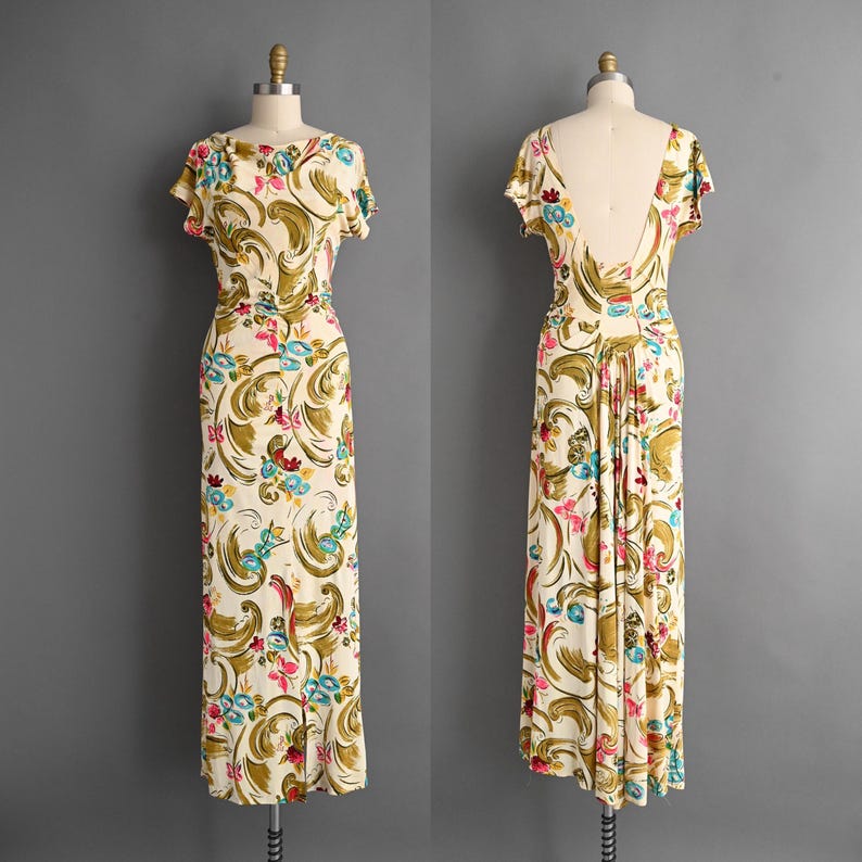 May include: A vintage cream-colored dress with a colorful floral pattern. The dress features a scoop neckline, short sleeves, and a fitted waist. The back of the dress has a low, open design. The floral pattern includes shades of green, pink, and blue.