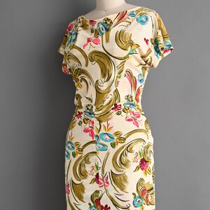 May include: Cream-colored vintage dress with a floral pattern. The dress features a scoop neckline, short sleeves, and a fitted silhouette. The floral design includes pink, blue, and yellow flowers with olive green accents. The dress is displayed on a mannequin.