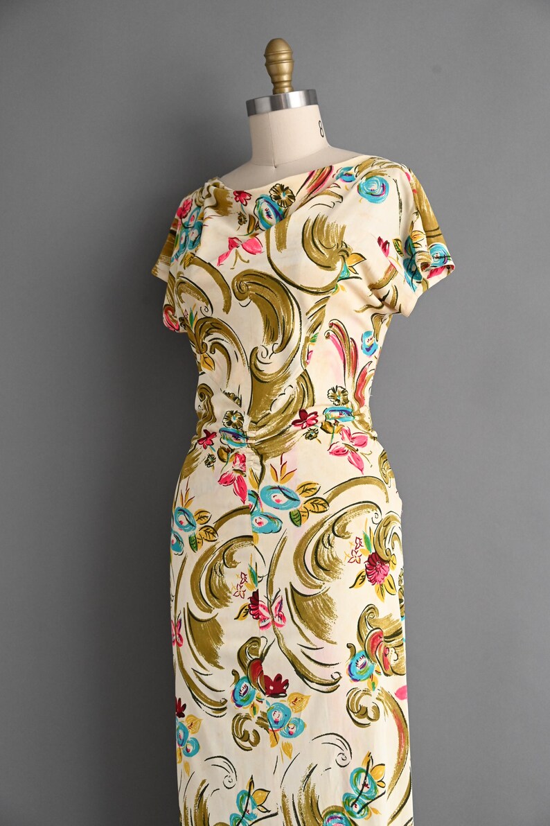 May include: Cream-colored vintage dress with a floral pattern. The dress features a scoop neckline, short sleeves, and a fitted silhouette. The floral design includes pink, blue, and yellow flowers with olive green accents. The dress is displayed on a mannequin.