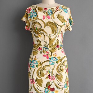 May include: A vintage cream-colored dress with a floral pattern. The dress features a scoop neckline, short sleeves, and a fitted waist. The floral design includes blue, pink, and red flowers with olive green and gold accents. The dress is displayed on a mannequin.