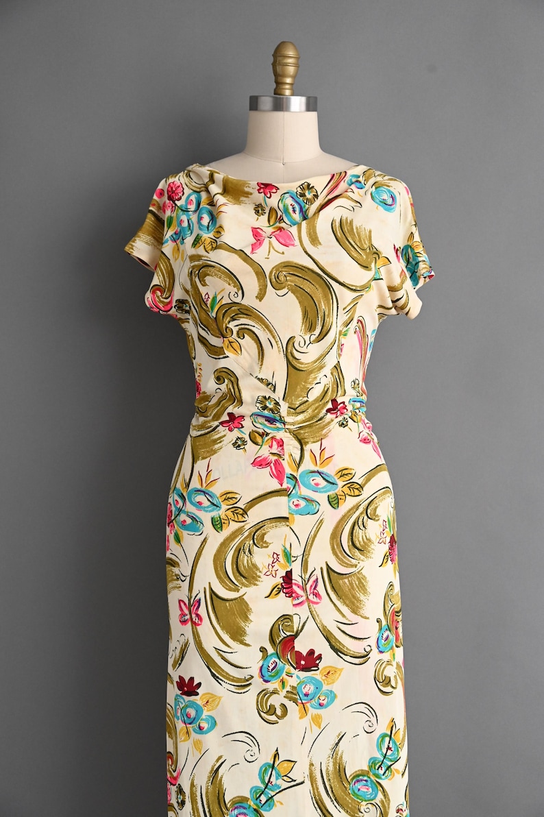 May include: A vintage cream-colored dress with a floral pattern. The dress features a scoop neckline, short sleeves, and a fitted waist. The floral design includes blue, pink, and red flowers with olive green and gold accents. The dress is displayed on a mannequin.
